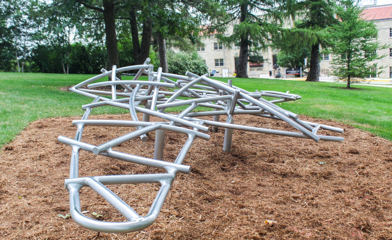 20 feet wide and 12 feet long aluminum sculpture with abstract, bird-like shape, standing in a grassy area with trees, building in the background.