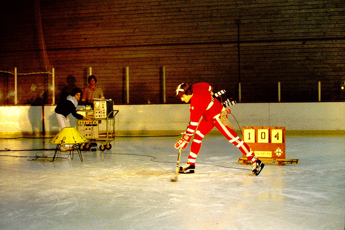 A hockey player shoots while two researchers look on.