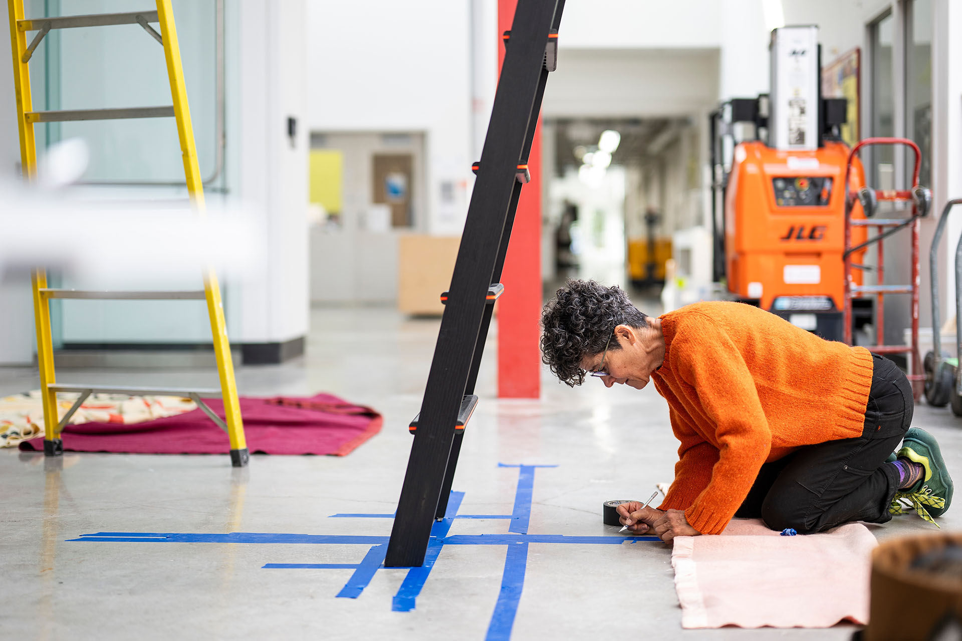 UW alumni artist Chele Isaac takes measurements on the floor amid blue painters tape in preparation to install her artwork