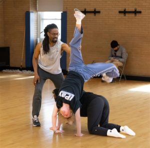 A student performs a handstand over another student, who is on all fours on the floor, while instructor Omari Carter stands to the side and offers hands-on support. 